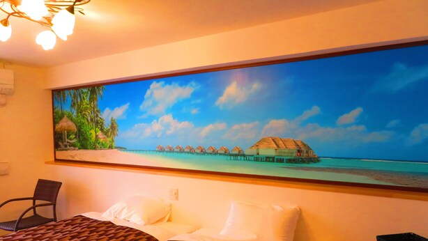 Une chambre d’hôtel avec un lit, une chaise, un tableau représentant une scène de plage et un luminaire au plafond.