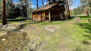 Metsäisellä alueella sijaitseva maalaishenkinen mökki, jonka ympärillä kohoaa korkeita mäntyjä.