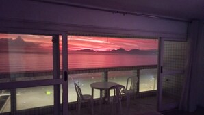 Un balcón con vista a un atardecer sobre el agua, una mesa con sillas y un vidrio.
