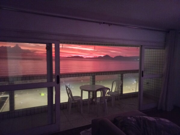 A balcony with a view of a sunset over water, a table with chairs, and a glass door.