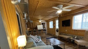 Ein gemütliches Blockhaus-Interieur mit Holzbalkendecke, einer Couch, einem Esstisch und einem an der Wand befestigten Fernseher.