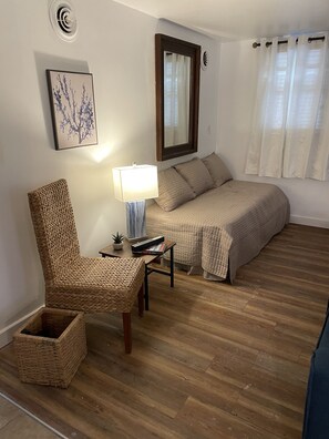 A cozy room with a bed, a chair, a small table with a lamp, a framed picture, and a window with curtains.