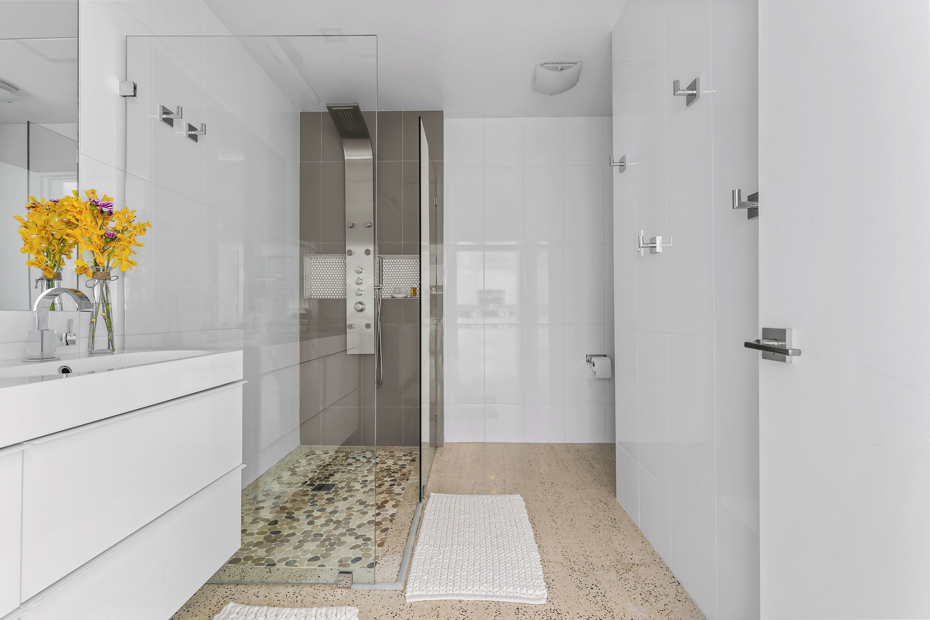 Un baño moderno con ducha envuelta en vidrio, tocador blanco y un jarrón decorativo con flores.