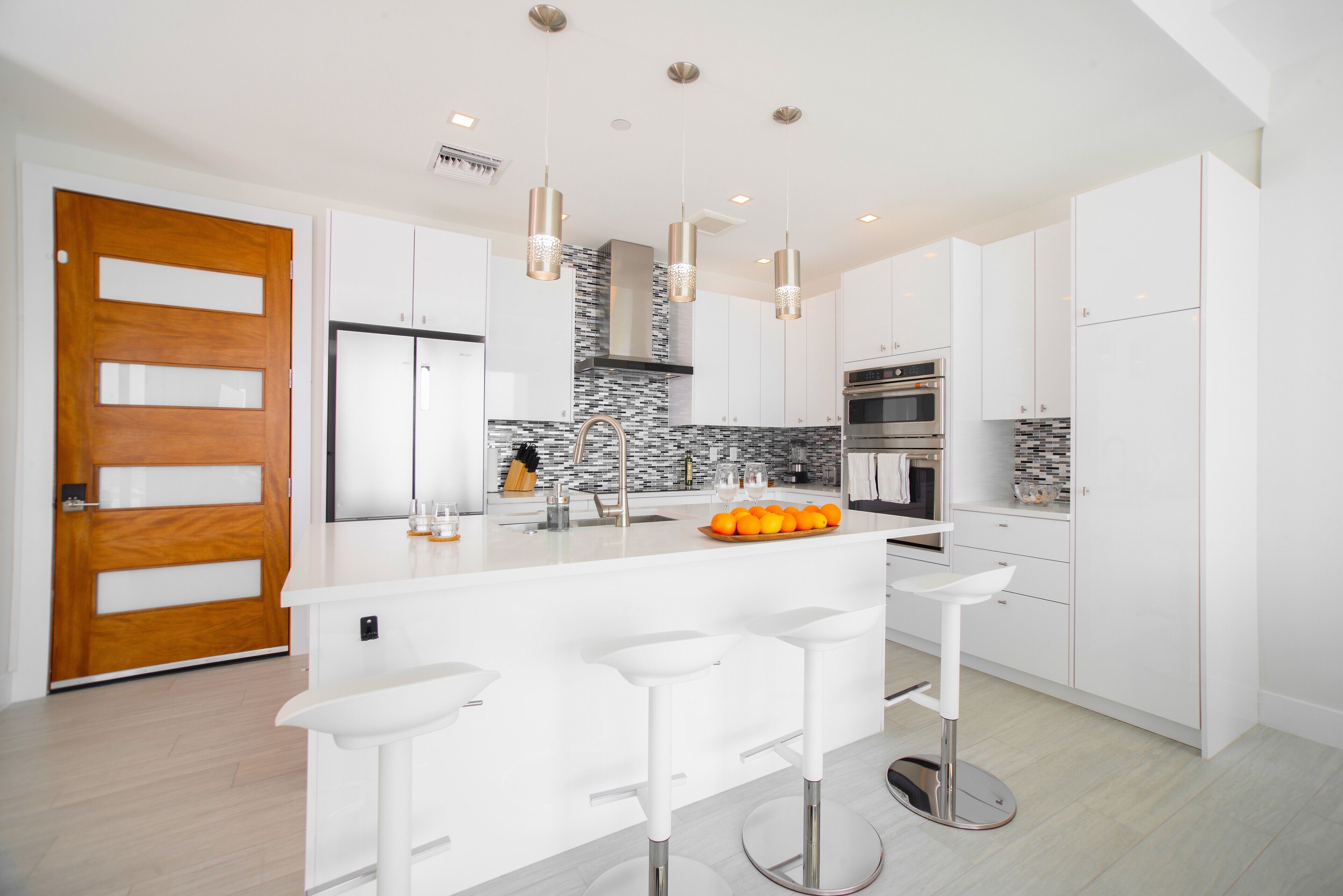 A modern kitchen with white cabinets, a central island with bar stools, a wooden door, and a stone backsplash.