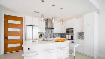 A modern kitchen with white cabinets, a central island with bar stools, a wooden door, and a stone backsplash.