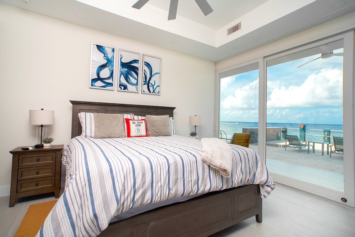 A bedroom with a large bed, a nightstand, a lamp, and a view of the ocean.