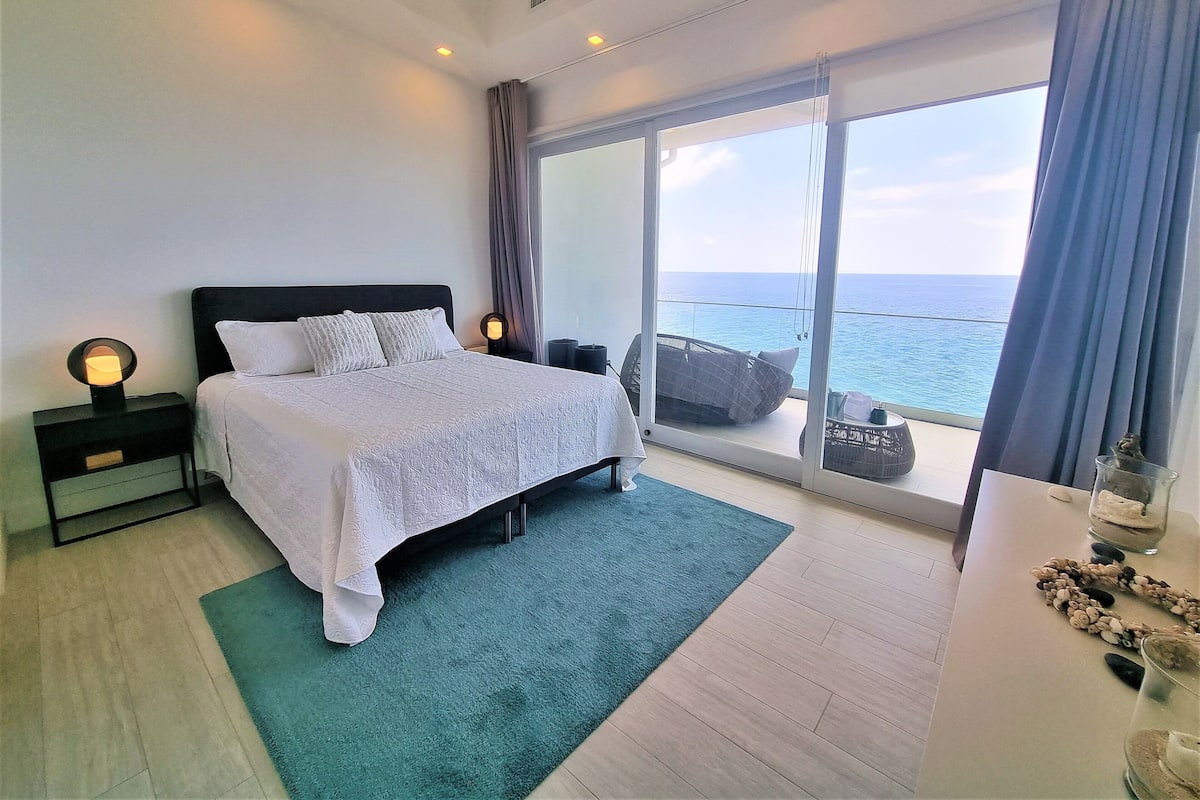 A modern bedroom with a large bed, a nightstand, a bedside lamp, a view of the sea through a sliding glass door, and a balcony with a chair.