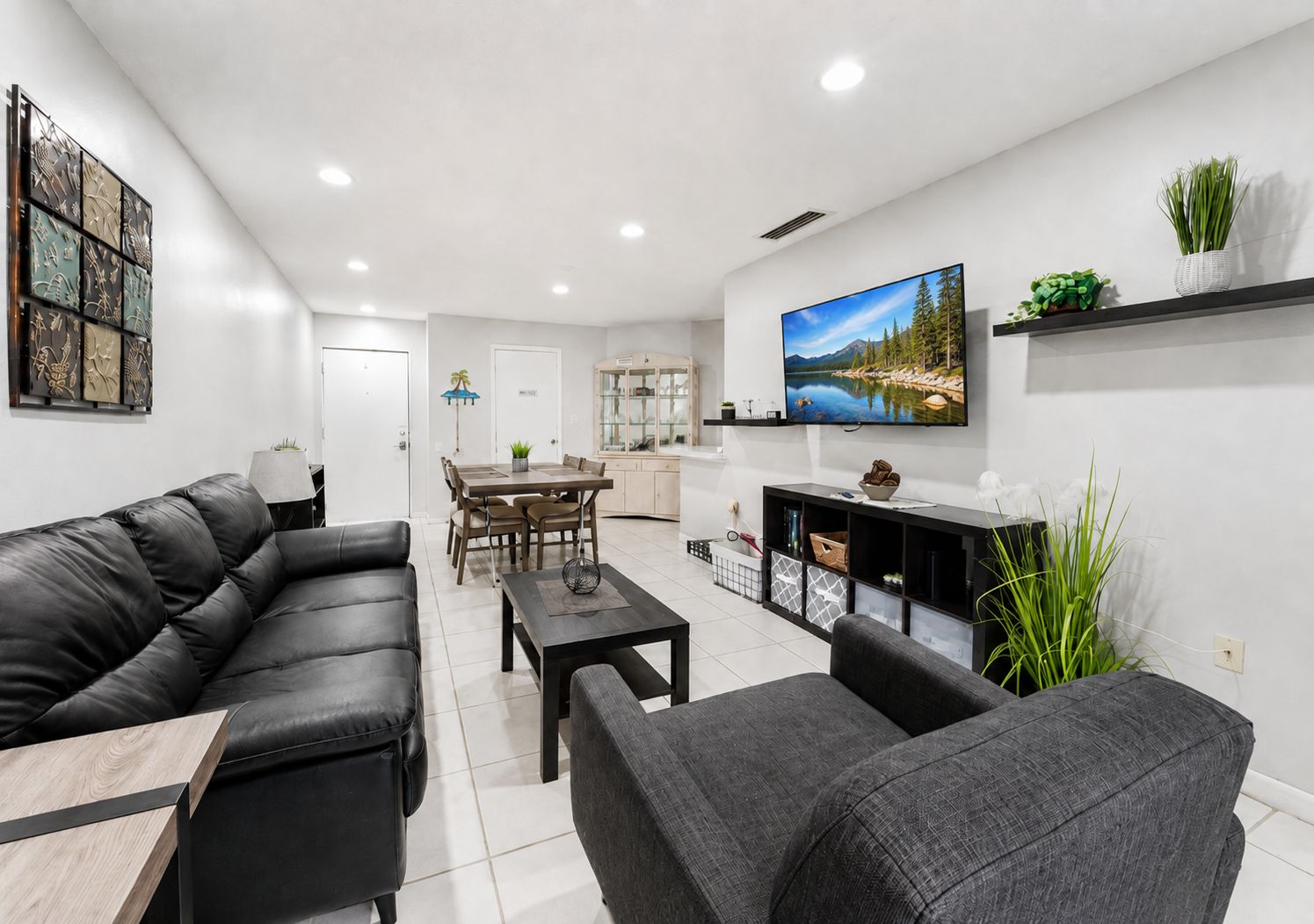 A modern living room with a black leather sofa, a grey armchair, a wooden coffee table, and a flat-screen TV mounted on the wall.