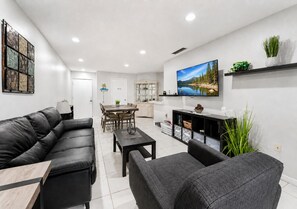 A modern living room with a black leather sofa, a grey armchair, a wooden coffee table, and a flat-screen TV mounted on the wall.