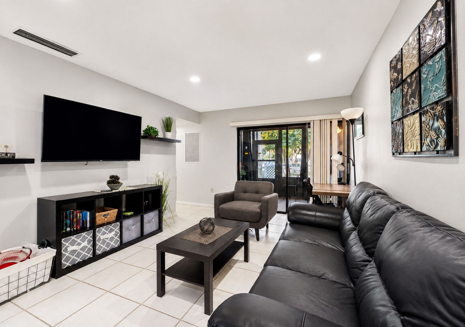 A modern living room with a flat-screen TV, a black leather sofa, a grey armchair, a coffee table, and a bookshelf.