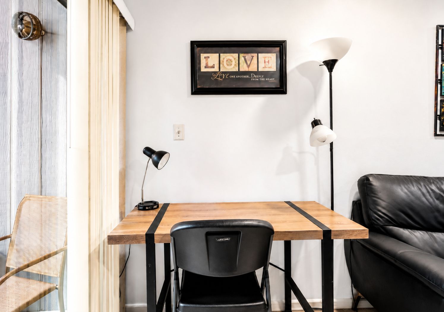 Een kamer met een houten tafel, een zwarte stoel, een zwarte vloerlamp, een witte lamp en een ingelijst citaat aan de muur.