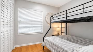 Quarto com beliche, janela com persianas, mesa de cabeceira com abajur e relógio na parede.