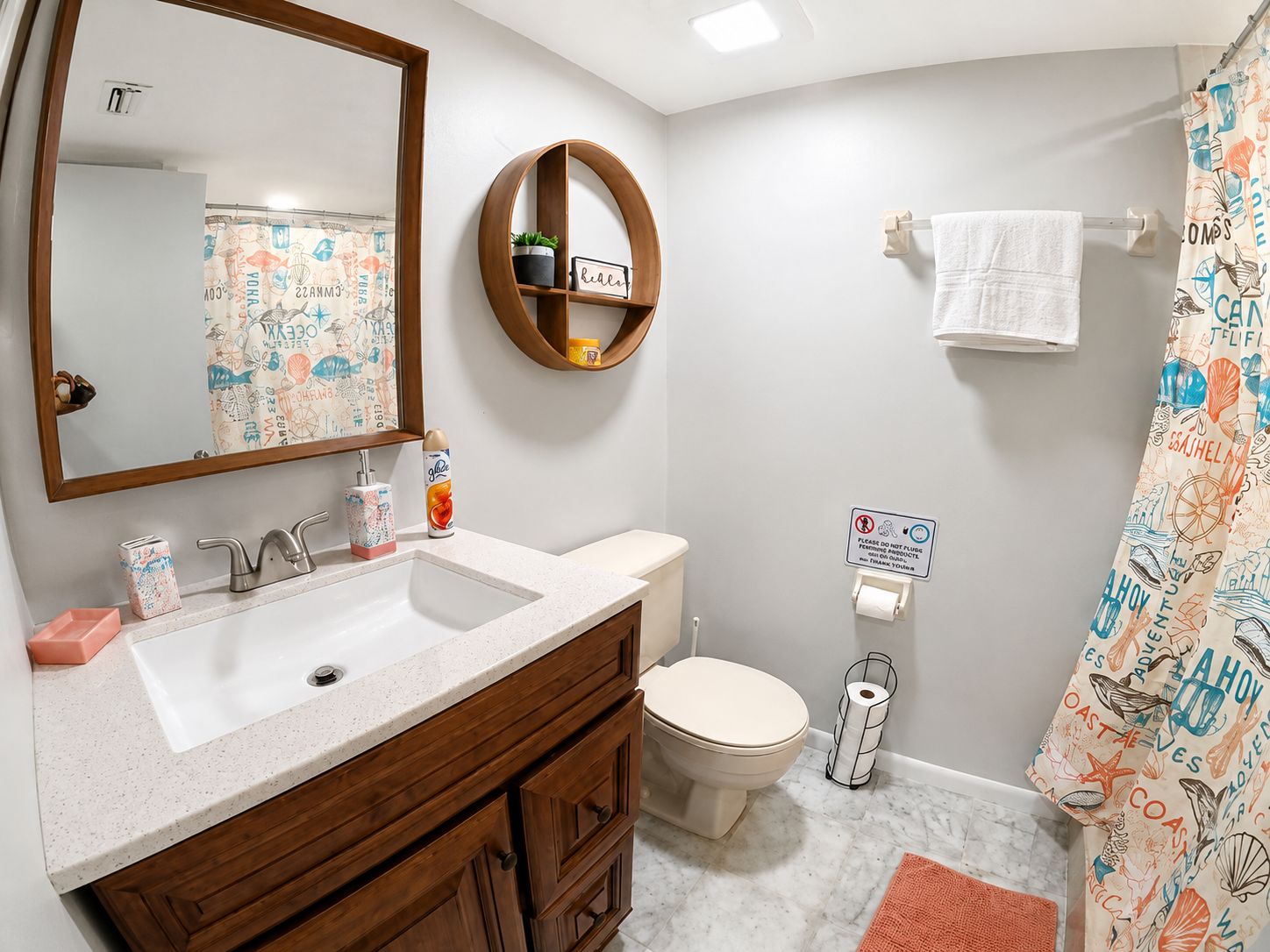 A bathroom with a wooden vanity, a toilet, a shower curtain with nautical prints, and a sign on the wall.