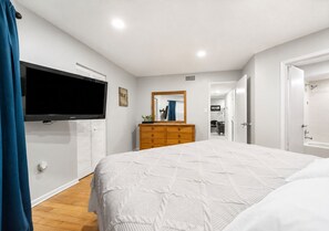 Quarto com uma cama grande, cômoda, televisão e porta que dá para outro espaço.