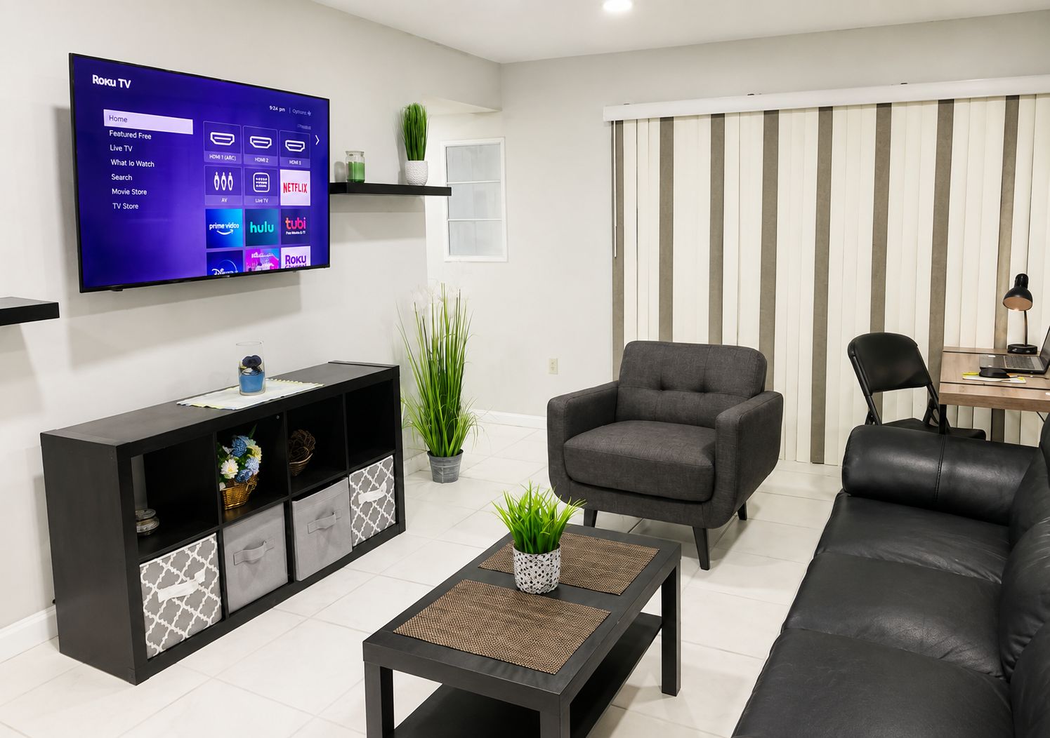 Een moderne woonkamer met een flatscreen televisie, een zwarte bank, een grijze fauteuil, een salontafel en een zwart tv-meubel met decoratieve objecten.