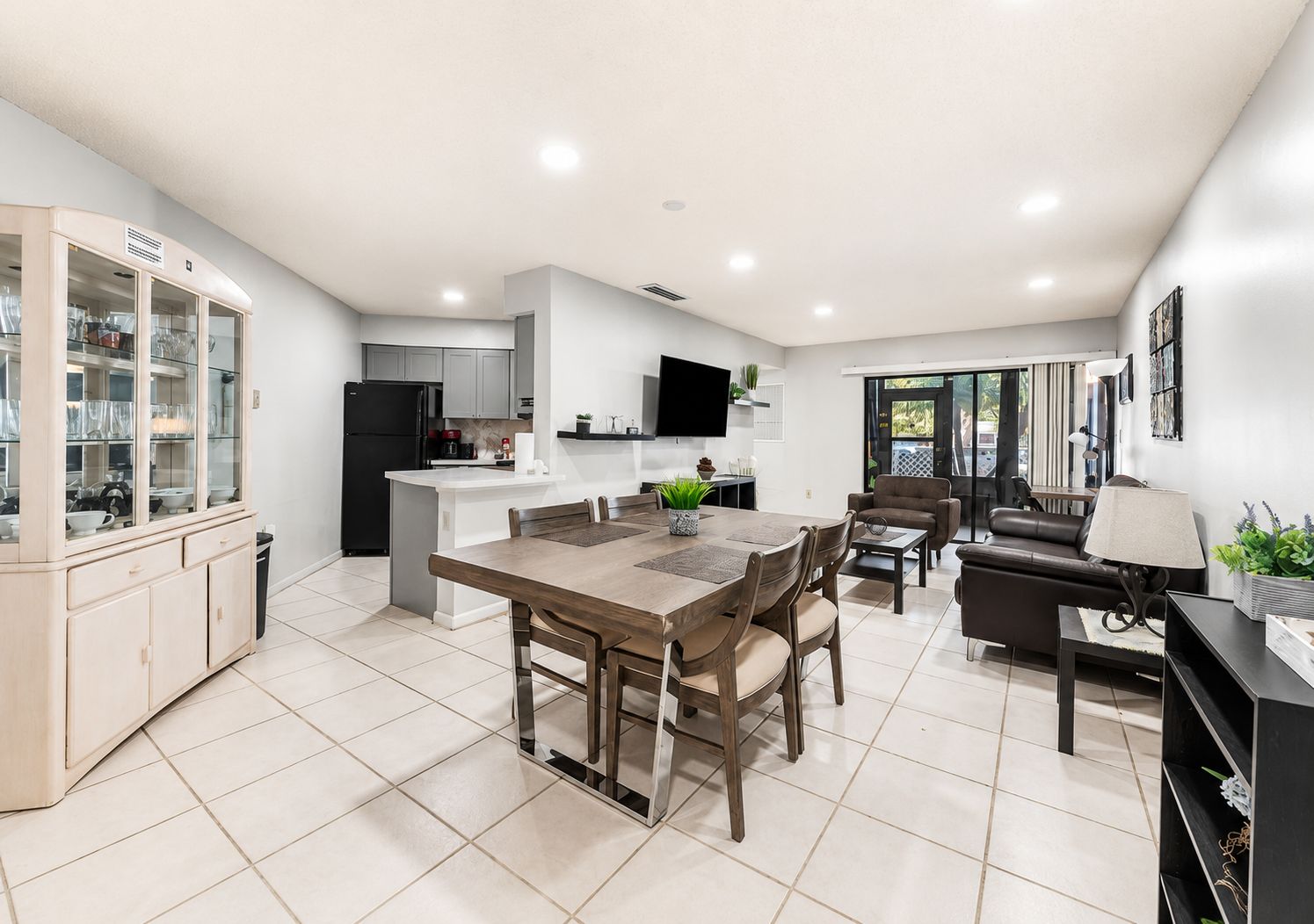 A modern kitchen with a dining area, a living room with a sofa and a TV, and a sliding glass door leading to an outdoor area.
