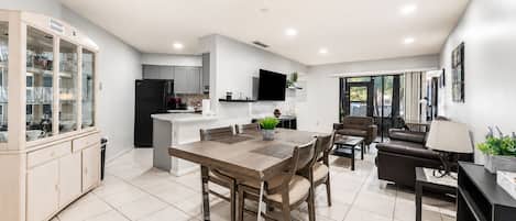 A modern kitchen with a dining area, a living room with a sofa and a TV, and a sliding glass door leading to an outdoor area.
