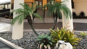 Una entrada de hotel con un porche sostenido por columnas, una escalera que lleva a la entrada y una zona ajardinada con plantas y rocas.