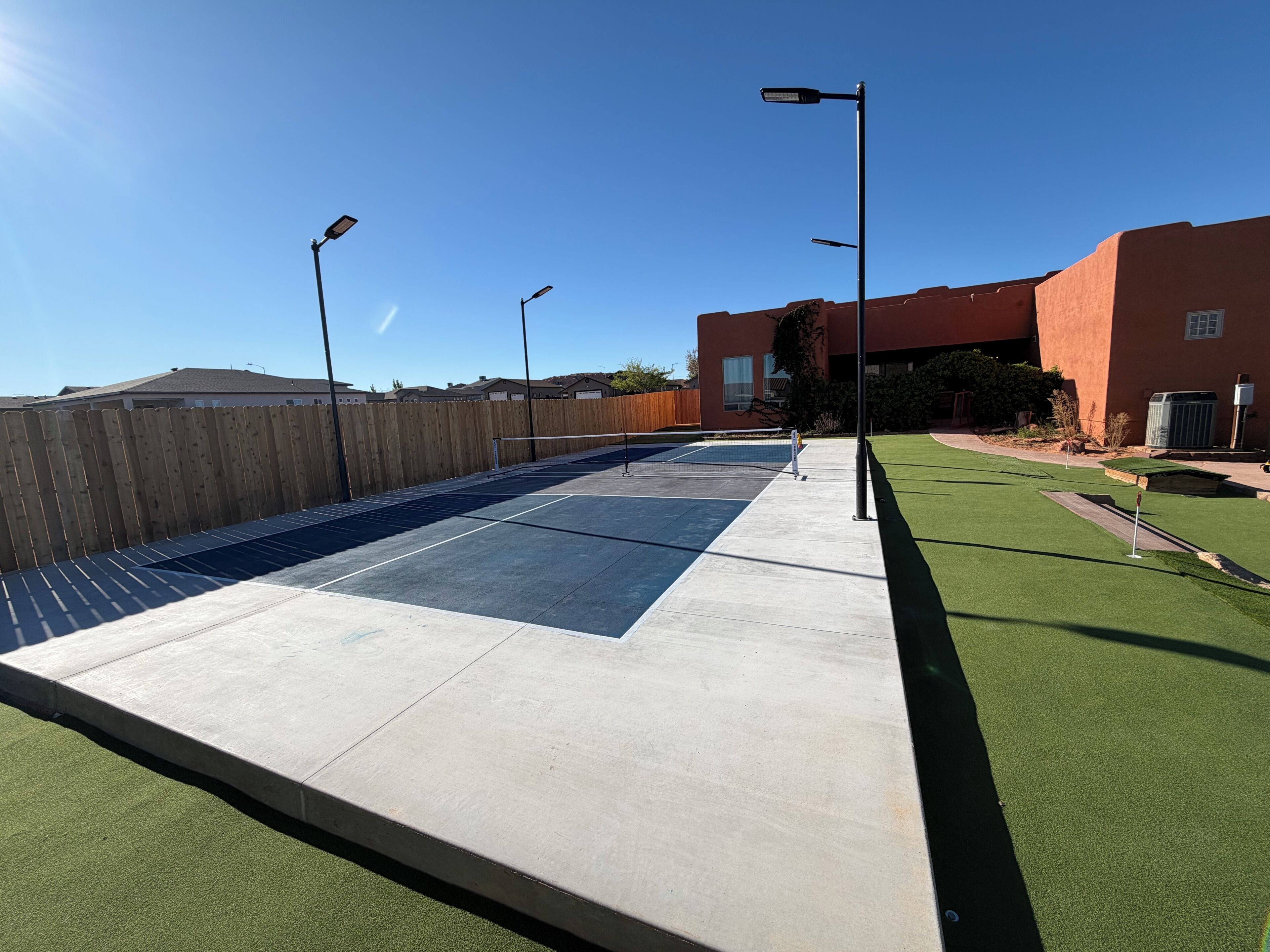 A tennis court with a wooden fence, street lamps, and a building in the background.