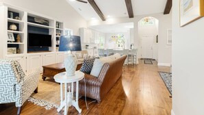A spacious living room with a wooden floor, a sofa, a coffee table, and a TV. There is a kitchen area with white cabinets and a dining table. - Pet Friendly Long-Term Rental - 25 Day Min  - Fenced Backyard - Grill (St. James)