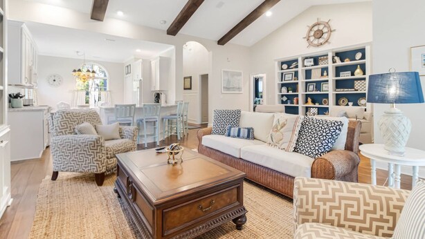 A spacious living room with a high ceiling, wooden beams, a large sofa, patterned armchairs, a coffee table, and a bookshelf filled with decorative items. - Pet Friendly Long-Term Rental - 25 Day Min  - Fenced Backyard - Grill (St. James)