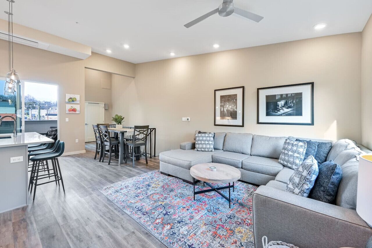 A modern living room with a grey sofa, a coffee table, and a patterned rug. There is a dining area with a table and chairs, and a kitchen area with a bar counter and stools.