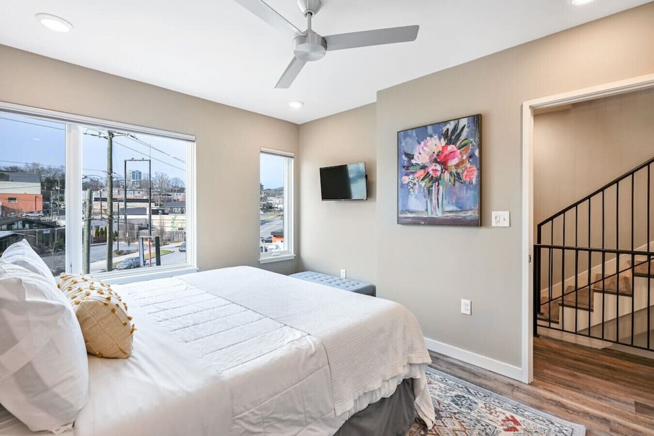 A bedroom with a large bed, a ceiling fan, a painting, and a view of the outside.