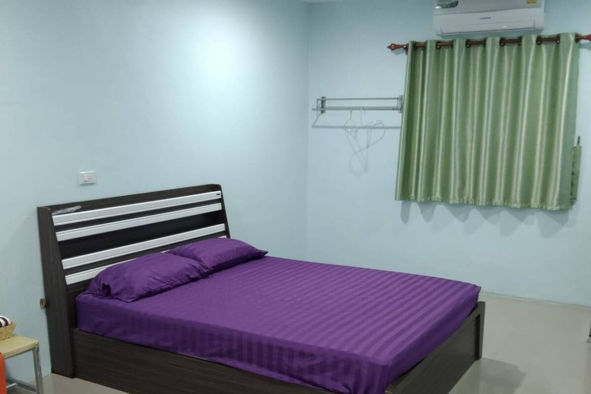 A bedroom with a bed, a nightstand, a small table, a wall-mounted air conditioner, and a window with curtains.