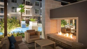 A modern outdoor lounge area with wicker furniture, a built-in fire feature, and a view of a swimming pool and adjacent buildings. - New! Master Suite w/ Skyline Views - Art Dist (Houston)