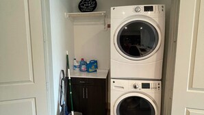 A laundry room with two stacked front-loading washing machines, a drying rack, and a shelf with cleaning supplies. - New! Master Suite w/ Skyline Views - Art Dist (Houston)