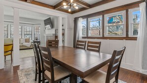 Un coin repas avec une table et des chaises en bois, de grandes fenêtres avec des rideaux et une vue sur l’extérieur.