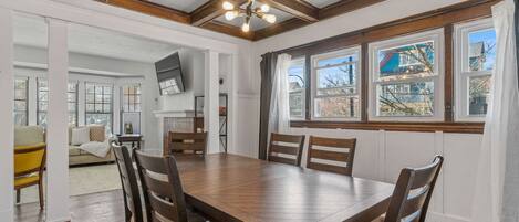 A dining area with a wooden table and chairs, large windows with curtains, and a view of the outside.