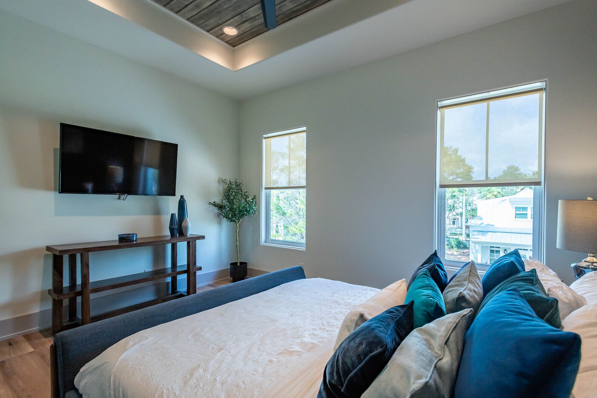 A modern bedroom with a large bed, a TV mounted on the wall, and two windows providing natural light.