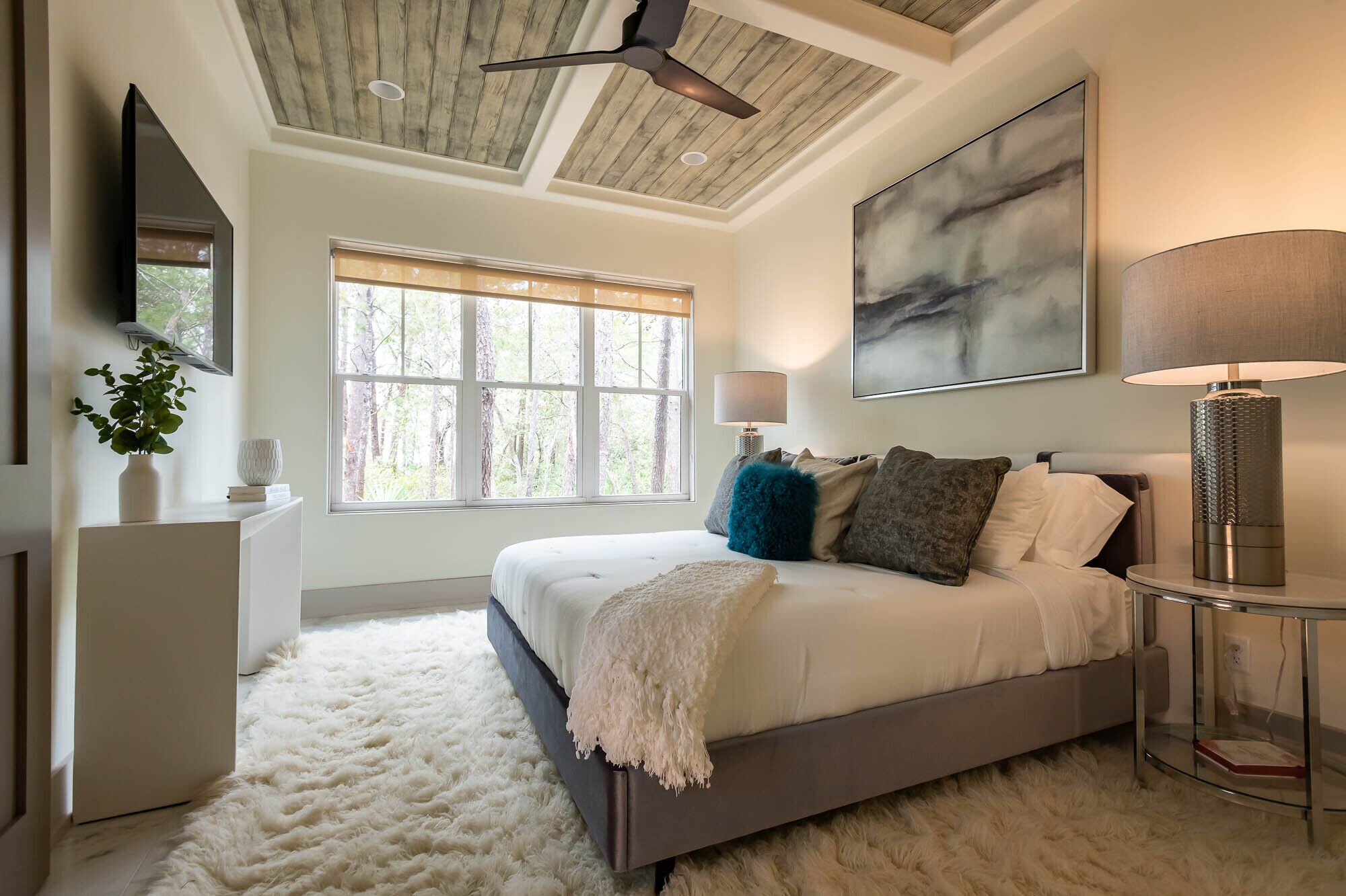 Ein Schlafzimmer mit einem großen Bett, einem Deckenventilator, einem Fenster mit Blick auf Bäume, einem Gemälde an der Wand und einer Lampe auf einem Nachttisch.