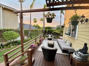 Una terraza de madera con mobiliario exterior, incluyendo un sofá y dos sillones, rodeada de vegetación y una casa al fondo.