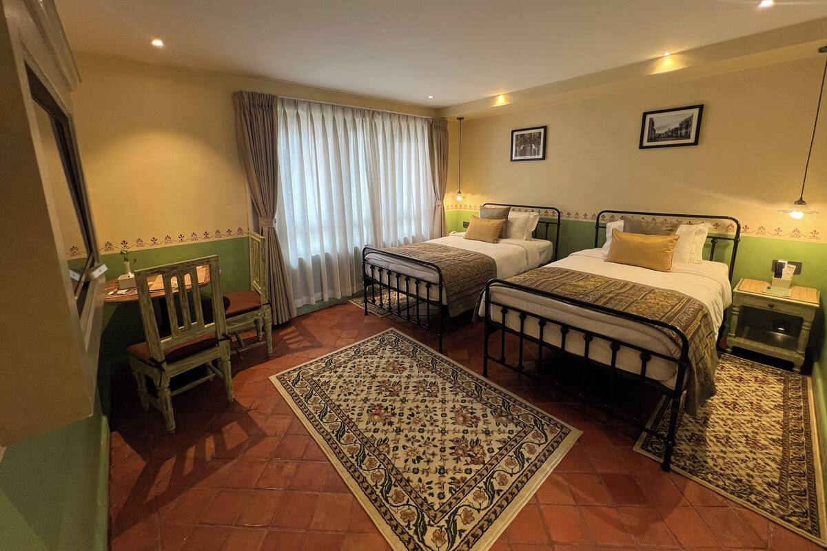 Une chambre avec deux lits, une chaise en bois, une petite table, un tapis à motifs et une fenêtre avec des rideaux.