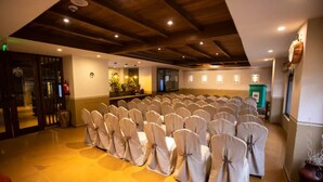 A conference room with rows of chairs covered in white chair covers, wooden ceiling beams, and a reception area with a counter and plants. - Seto Ghar Hotel Pvt. Ltd. (Kathmandu)