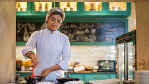 A chef in a white uniform cooking in a kitchen with teal cabinets and a chalkboard menu in the background. - Seto Ghar Hotel Pvt. Ltd. (Kathmandu)