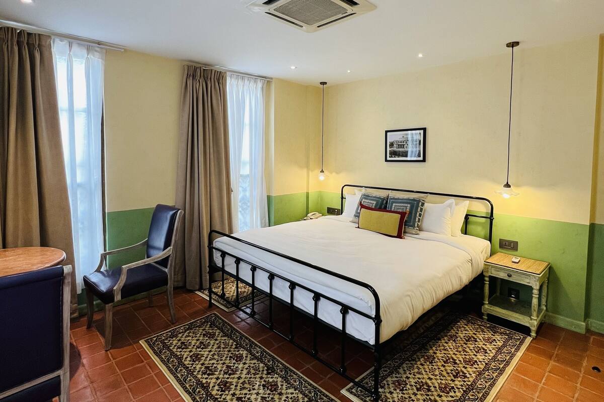 Une chambre à coucher avec un lit, une chaise, une table de chevet, une lampe et une fenêtre avec des rideaux.