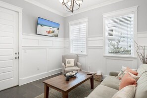 A living room with a wooden coffee table, a beige sofa, and a chair with a cushion. There is a painting on the wall and a window with blinds.