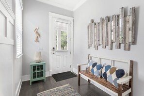 Een gang met een witte deur, een blauwgroene kast, een houten bank met gestreepte kussens, en een spiegel aan de muur.