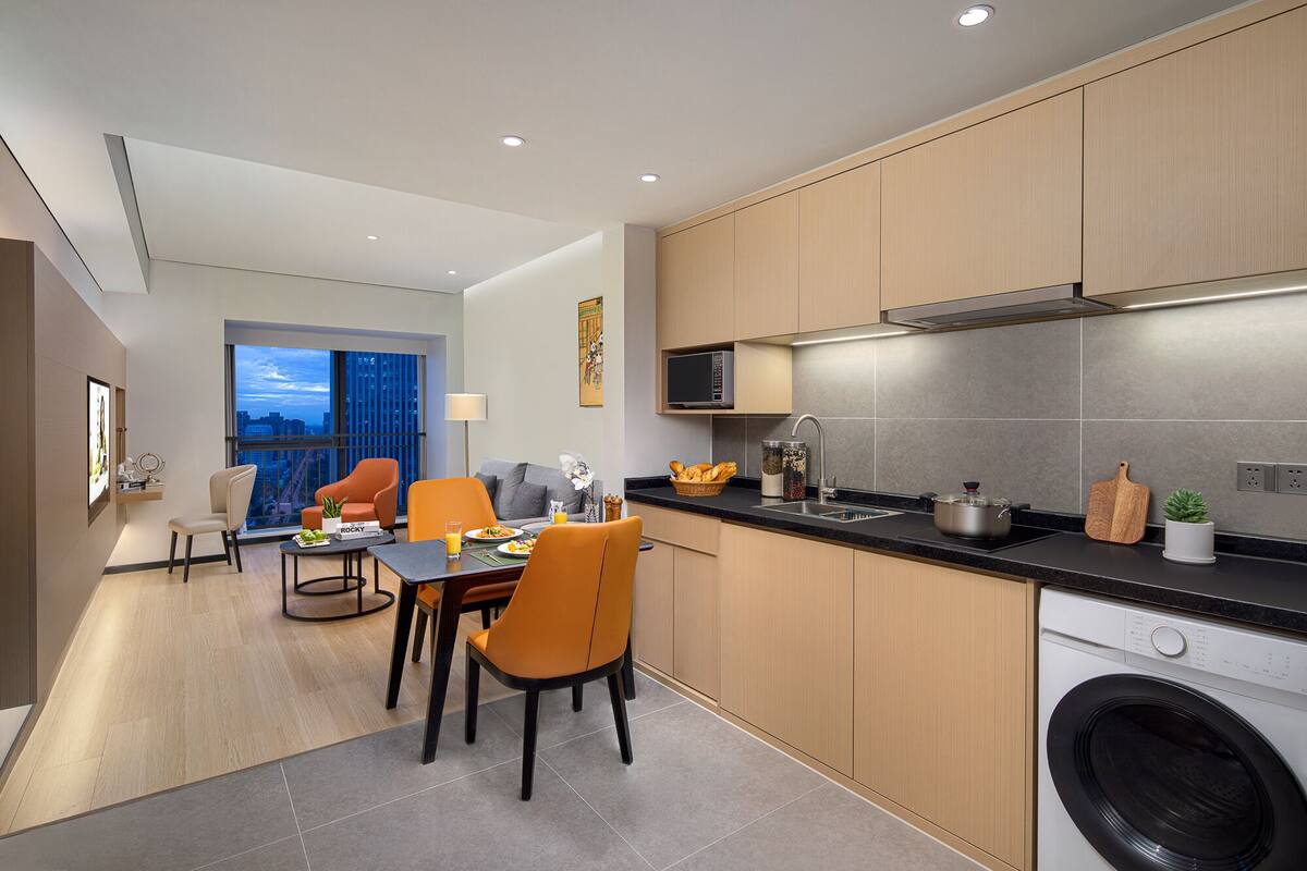 A modern kitchen with a built-in microwave, a sink, and a washing machine. There is a dining area with orange chairs and a table, and a large window offering a city view.
