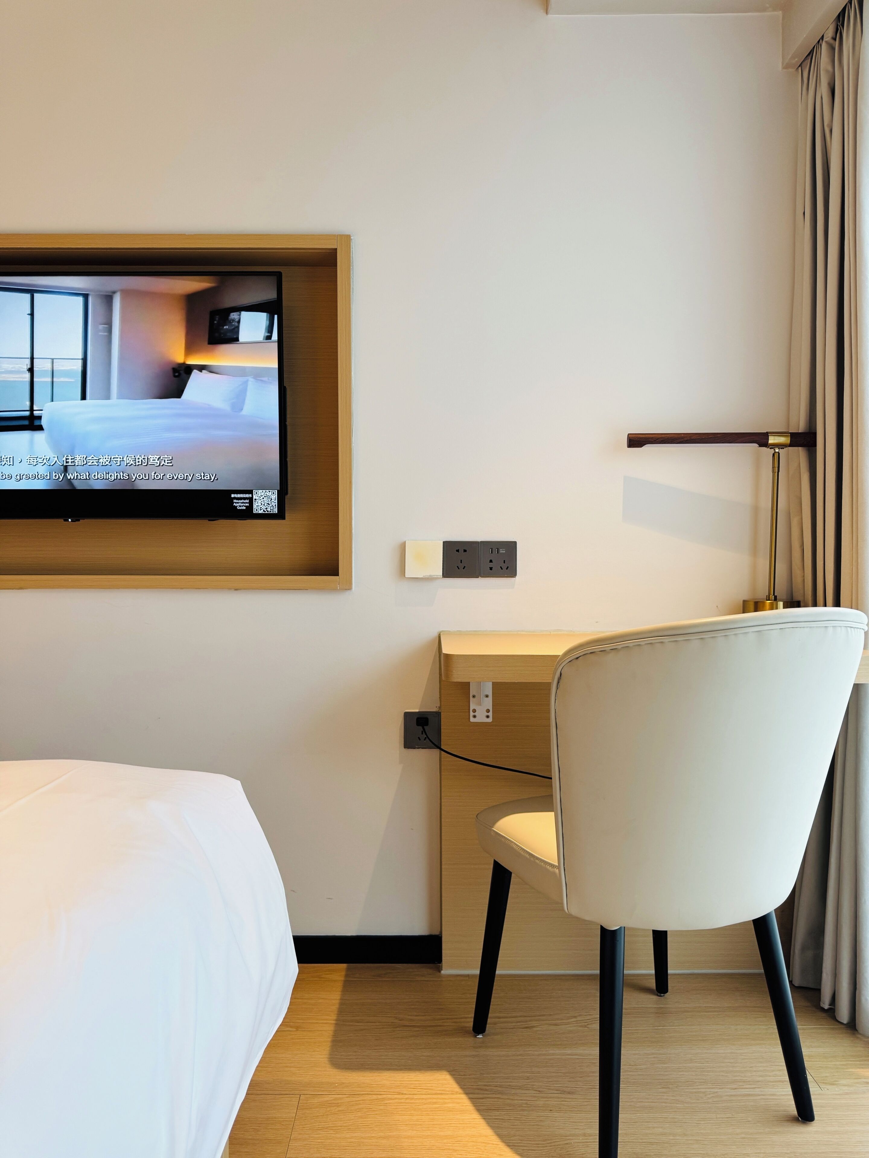 Ett hotellrum med en säng, ett skrivbord, en stol, en tv som är monterad på väggen och en gardinstång.