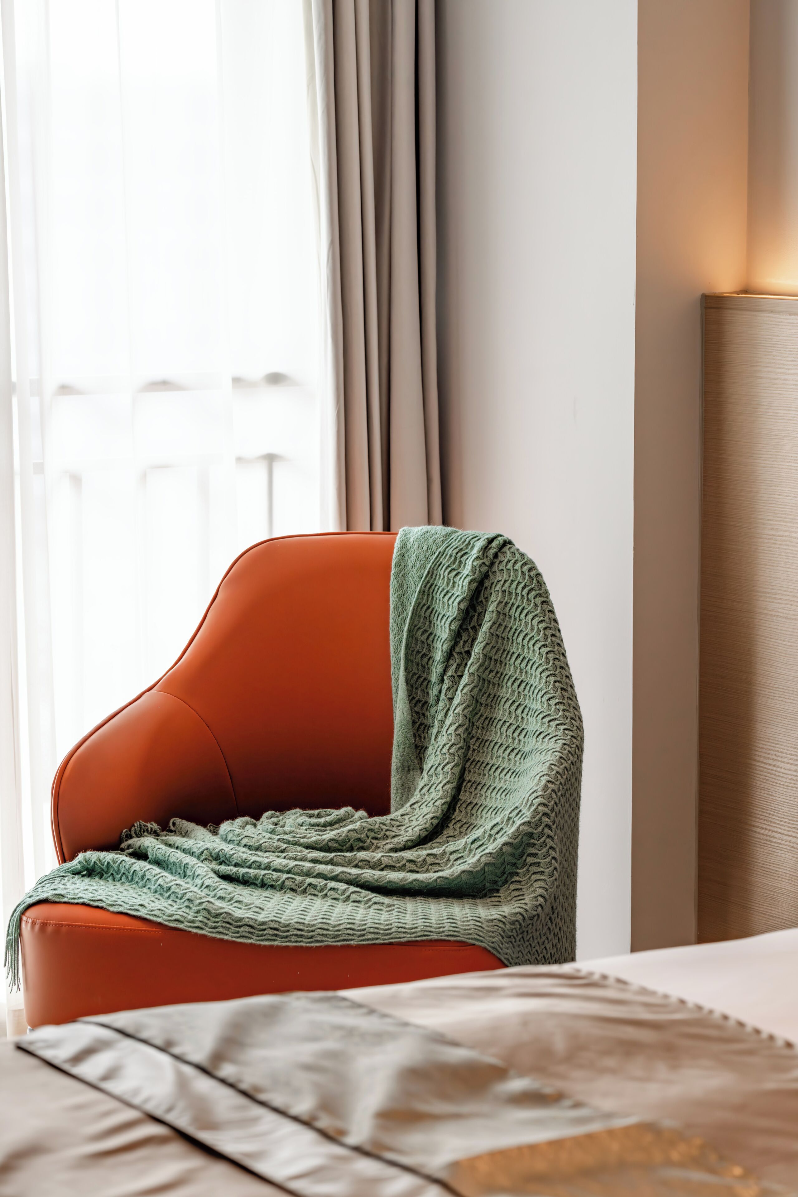 Una habitación con una silla naranja, un plaid verde de punto, una cama con una manta beige y una lámpara de noche.