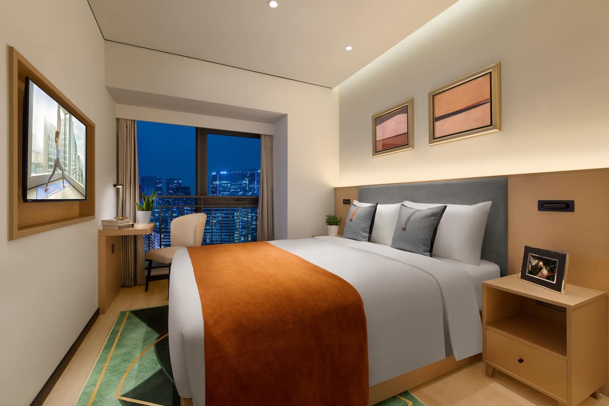 A modern hotel room with a large bed, a desk, a chair, and a view of the cityscape at night.