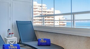 Un balcón con una silla, una mesita con adornos y vistas a edificios y al mar.