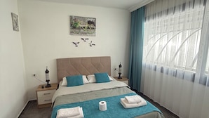 A neatly arranged bedroom with a bed, bedside tables, a hanging light fixture, a wall-mounted artwork, and a window with curtains. - Arslan Homes1  (Marmaris)