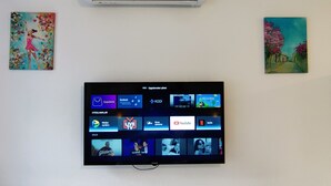 A wall-mounted air conditioner, a flat-screen TV displaying various streaming services, and two framed artworks.
