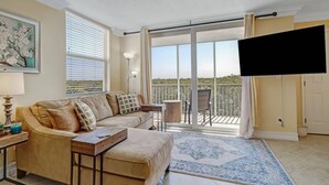 A living room with a beige sofa, a coffee table, a TV mounted on the wall, and a balcony with a view of greenery. - IN-UNIT WASHER/DRYER | WALK-IN CLOSET | FREE WIFI | KING BED + QUEEN SLEEPER | STEPS TO BONITA BEACH (Bonita Springs)