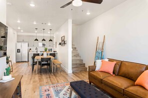 Una cocina moderna con armarios blancos, una zona de comedor con una mesa y sillas de madera, un sofá de cuero marrón y una escalera que sube a otro nivel.